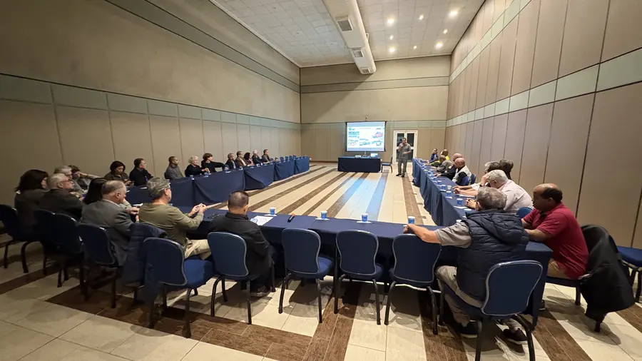 Sala de reunião com participantes nas mesas em formato de U acompanhando apresentação