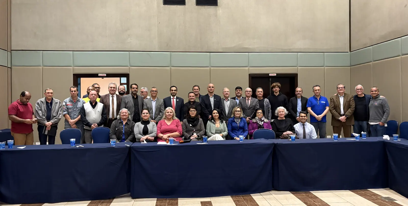 Foto oficial de reunião do Movimento Guarujá 2034: cerca de cinquenta participantes em pé atrás de mesas em formato de U