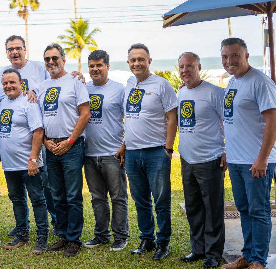 Integrantes do Movimento Guarujá 2034, com camisetas do Movimento, reunidos ao ar livre com palmeiras e o mar ao fundo